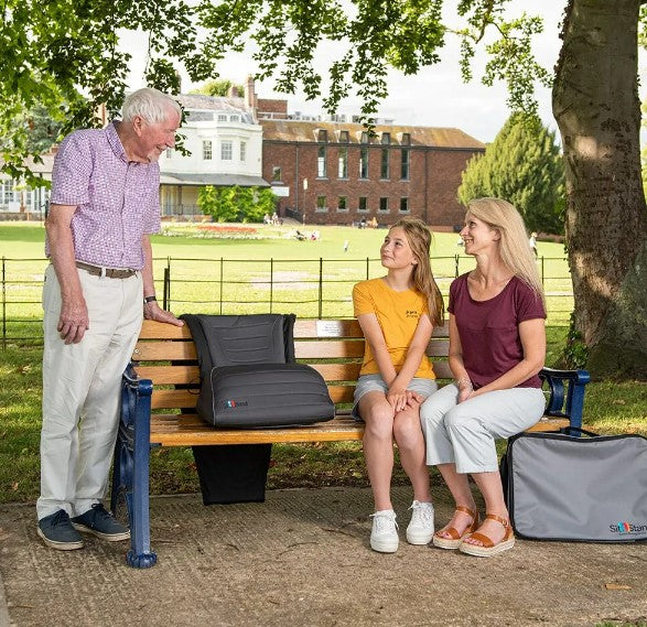 An older man stands by a park bench, smiling at two women sitting on the bench, next to large gray portable seat cushions, with a grassy area and buildings in the background.