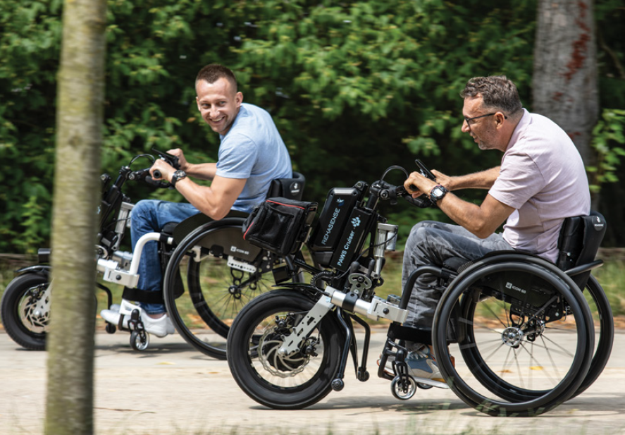 Two men in wheelchairs with front-mounted hand-bike attachments ride outdoors on a sunny day, smiling and enjoying the activity, with trees in the background.