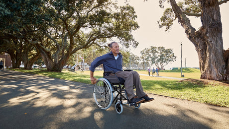 A man in a wheelchair moves along a paved path through a sunny park with large trees and grassy areas. A couple walks in the background.
