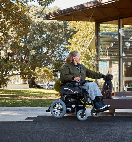 A person in a green jacket and gray skirt smiles while using a motorized wheelchair on a paved path. They are near a modern building with wooden accents. Trees and people are in the background.