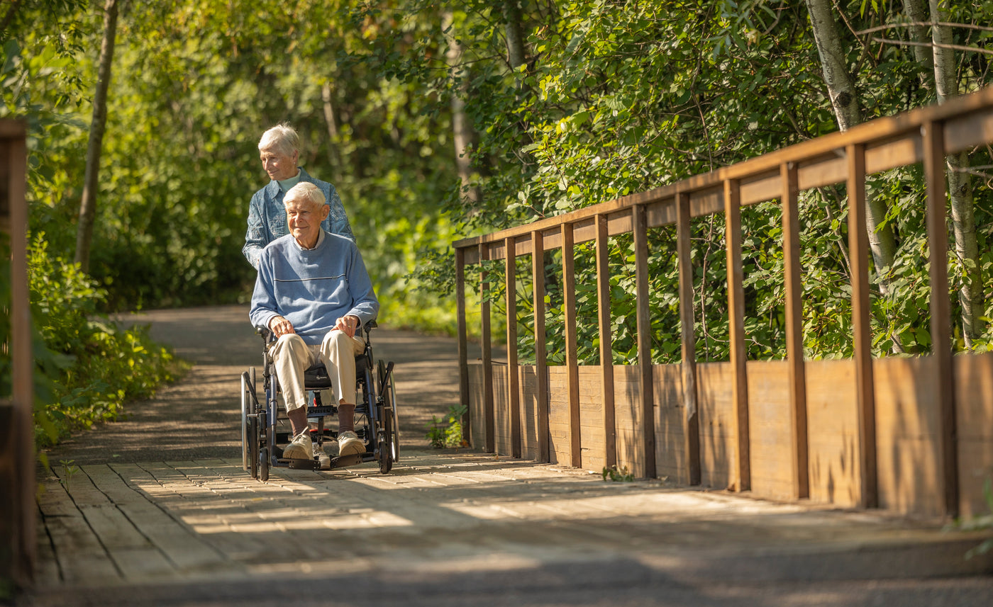 Woman pushing a man in a Ki Mobility Liberty wheelchair over a scenic bridge surrounded by lush trees, showcasing accessibility and mobility support.