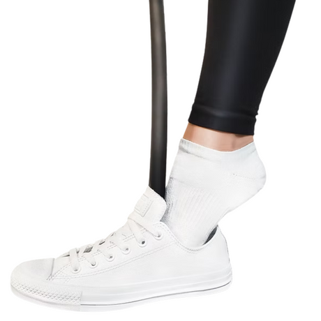 A person with limited mobility uses the ShoeStik Shoehorn by ShoeStik, featuring an ergonomic handle, to easily slip on white sneakers while wearing black leggings and white socks.